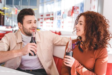 beautiful couple smiling and holding glass bottles with soda at cafe  clipart