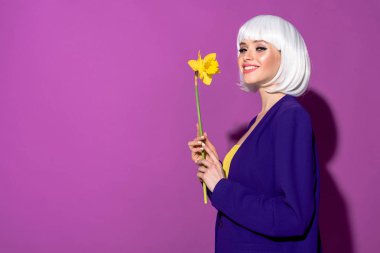 Inspired girl in white wig holding flower and smiling on purple background clipart