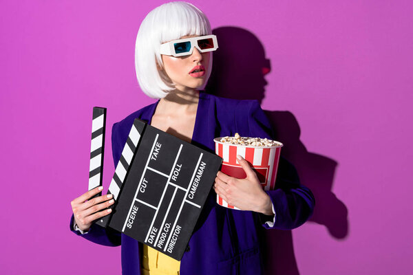Worried girl in 3d glasses holding clapperboard and popcorn on purple background