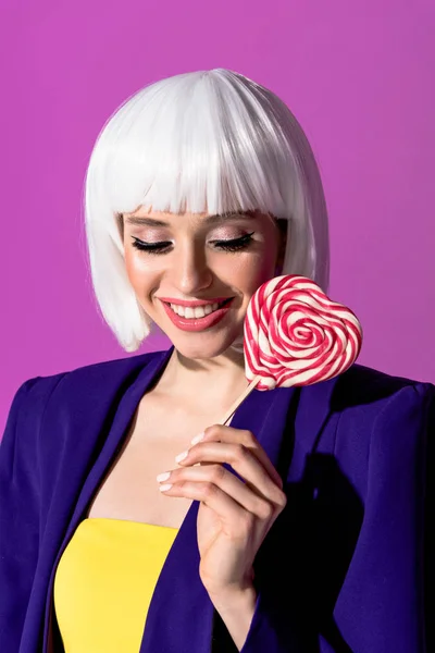 幸福的女孩在白色假发拿着心形棒棒糖查出在紫色 — 图库照片