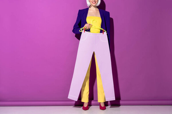 Cropped view of smiling woman holding hanger with paper pants on purple background