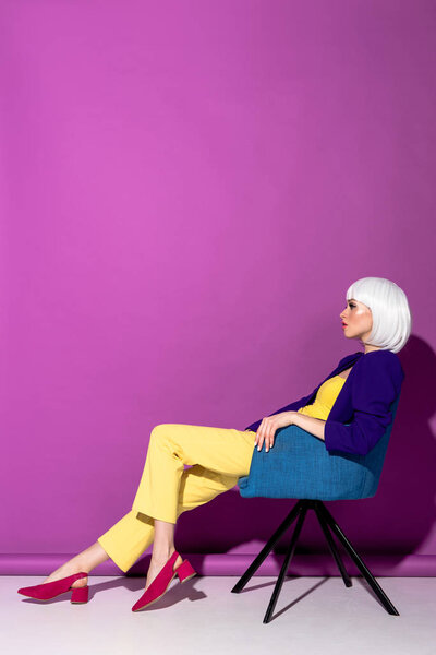 Side view of stylish girl in white wig sitting in armchair on purple background