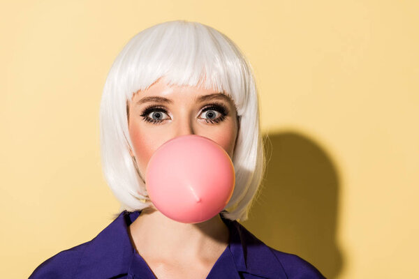 Amazed girl in white wig chewing bubble gum on yellow background
