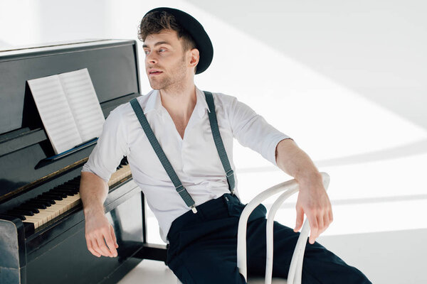 handsome pensive musician sitting on white chair and leaning on piano 