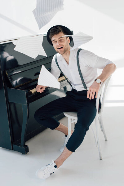 smiling musician in trendy clothing sitting by piano and throwing music book sheets 