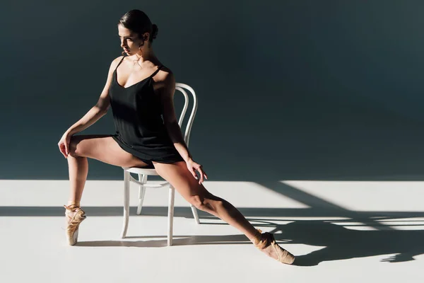 迷人的年轻芭蕾舞演员在黑色礼服伸展在白色的椅子上 — 图库照片
