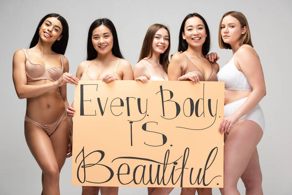 five smiling young multicultural women holding placard with "every body is beautiful" lettering isolated on grey