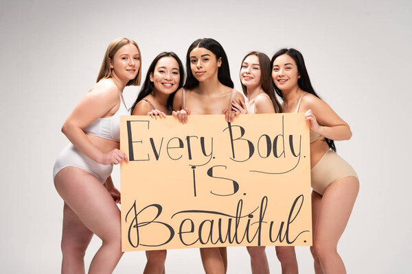 five smiling multiethnic girls holding placard with "every body is beautiful" lettering isolated on grey 