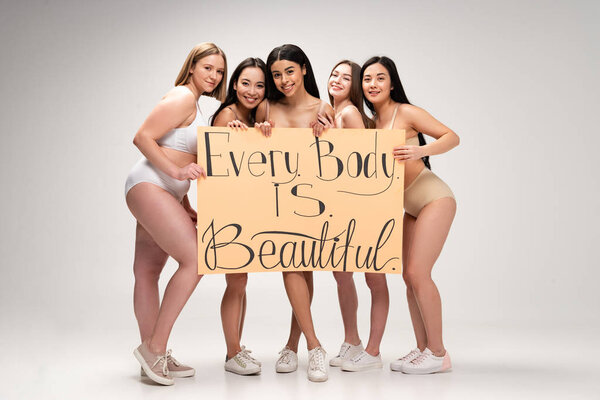 five pretty young multiethnic women in underwear and sneakers holding placard with "every body is beautiful" inscription 