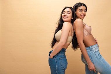 Mavi jeans ve sırt sırta duran ve bej, vücut pozitifliği kavramı izole kamera bakarak sutyen iki çok kültürlü mutlu kızlar