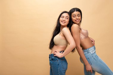 Mavi jeans ve sırt sırta duran ve gülümseyen sutyen iki oldukça çok kültürlü kızlar izole bej, vücut pozitifliği kavramı üzerinde