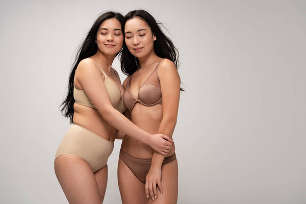 two beauty multiethnic young women with closed eyes in lingerie isolated on grey, body positivity concept