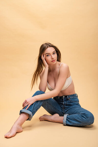 счастливая красивая девушка в синих джинсах и лифчике сидит и смотрит в камеру
