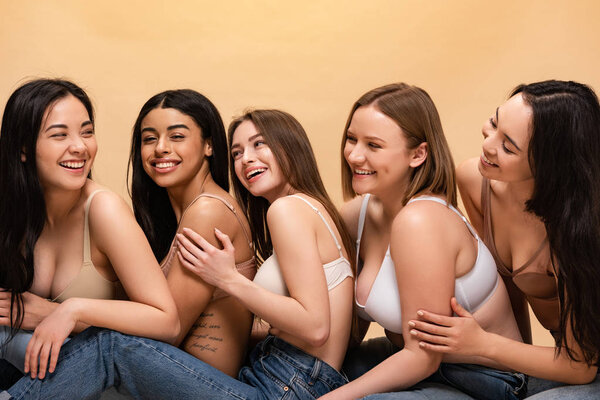 five cheerful multicultural girls sitting and leaning on each other isolated on beige, body positivity concept