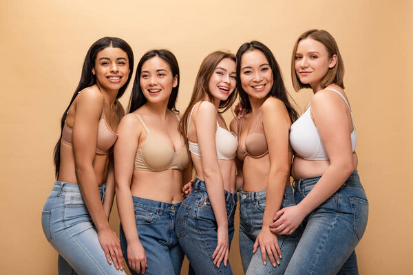 five beautiful multicultural girls smiling while posing at camera isolated on beige, body positivity concept