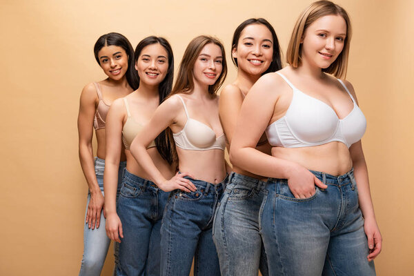 five pretty multicultural girls in blue jeans and bras smiling and looking at camera isolated on beige, body positivity concept