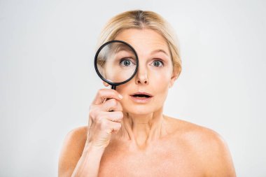 beautiful and surprised mature woman holding loupe and looking at camera on grey background  clipart