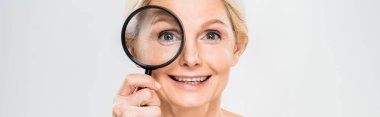 panoramic shot of beautiful and smiling mature woman holding loupe and looking at camera on grey background  clipart