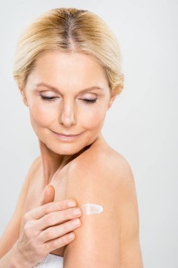 beautiful and mature woman looking down and applying cosmetic cream on shoulder isolated on grey 