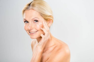 beautiful and mature woman looking at camera and applying cosmetic cream on face  clipart