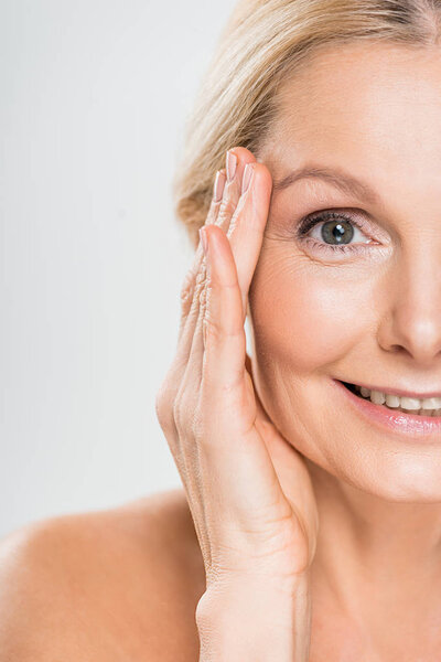 partial view of mature woman looking at camera and touching her face on isolated on grey 