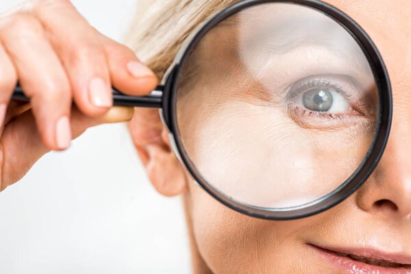 selective focus of mature woman looking at camera and holding loupe isolated on grey 