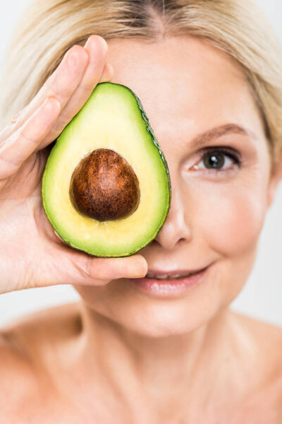 selective focus of beautiful and mature woman holding avocado and looking at camera isolated on grey