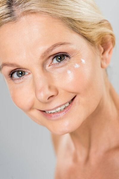 portrait of beautiful and mature woman looking at camera with cosmetic cream on face isolated on grey 