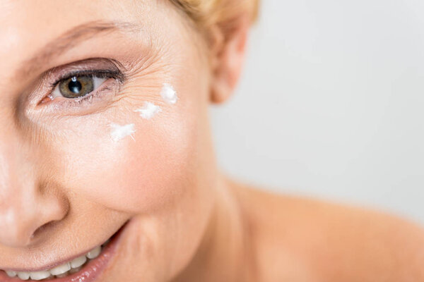 selective focus of beautiful and mature woman looking away with cosmetic cream on face isolated on grey 