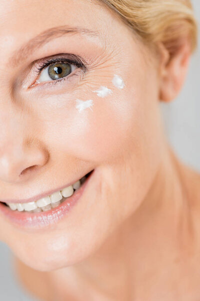 selective focus of beautiful and mature woman looking at camera with cosmetic cream on face isolated on grey 