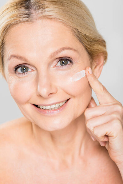 beautiful and mature woman looking at camera and applying cosmetic cream on face isolated on grey 