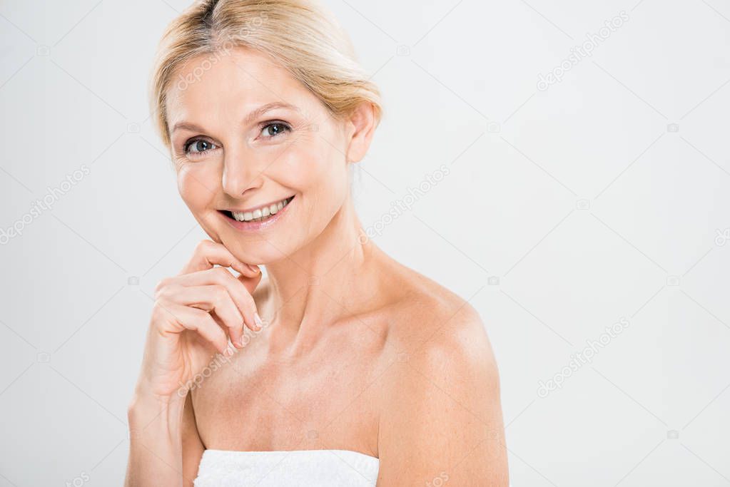 attractive and smiling mature woman looking at camera and touching her face on grey background 