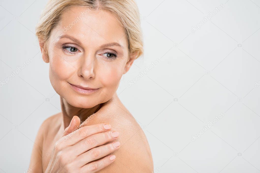 attractive and blonde mature woman looking away and touching her shoulder on grey background 