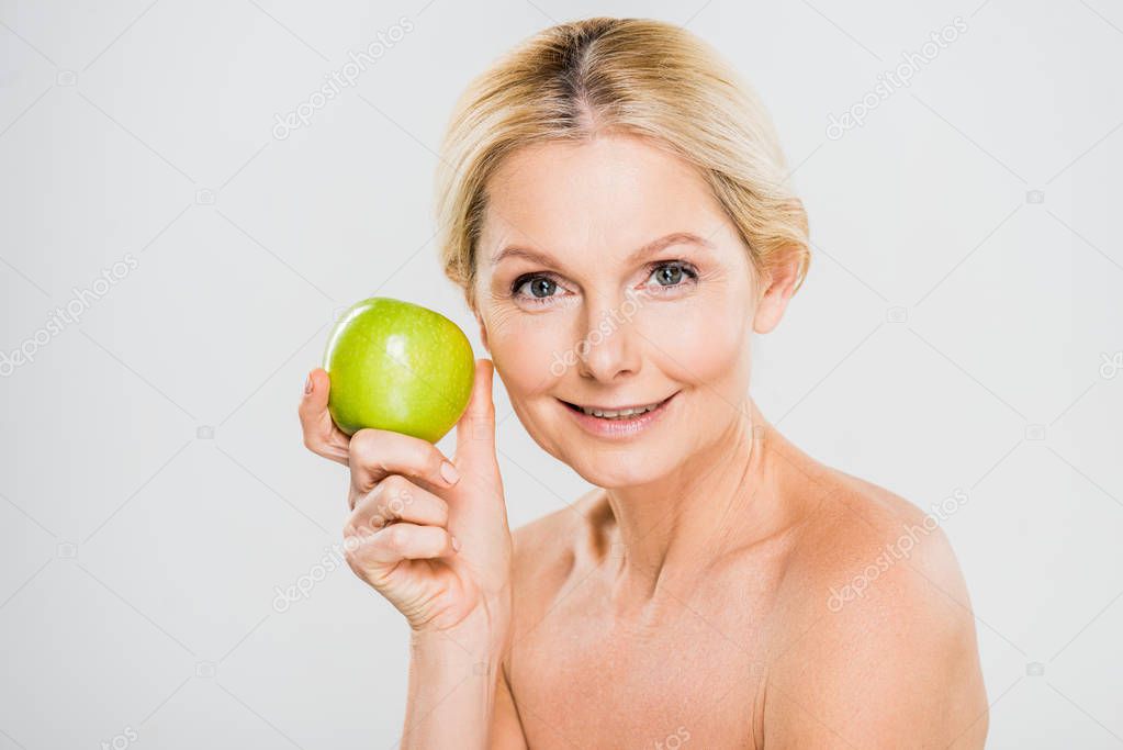 beautiful and smiling mature woman holding ripe apple and looking at camera on grey background 