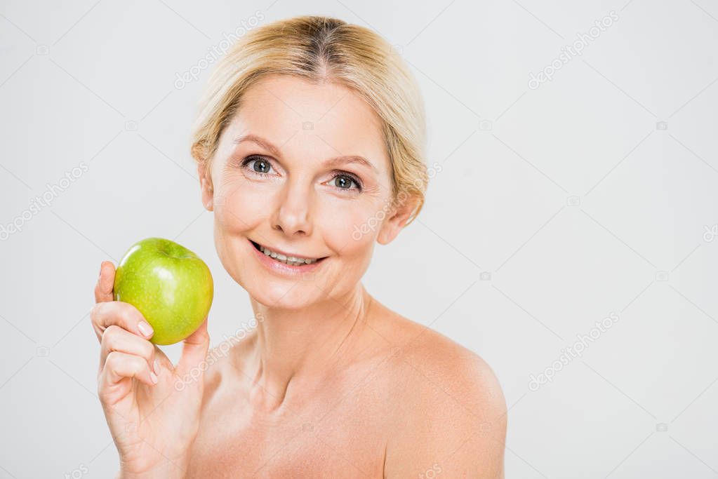 beautiful and smiling mature woman holding green apple and looking at camera on grey background 
