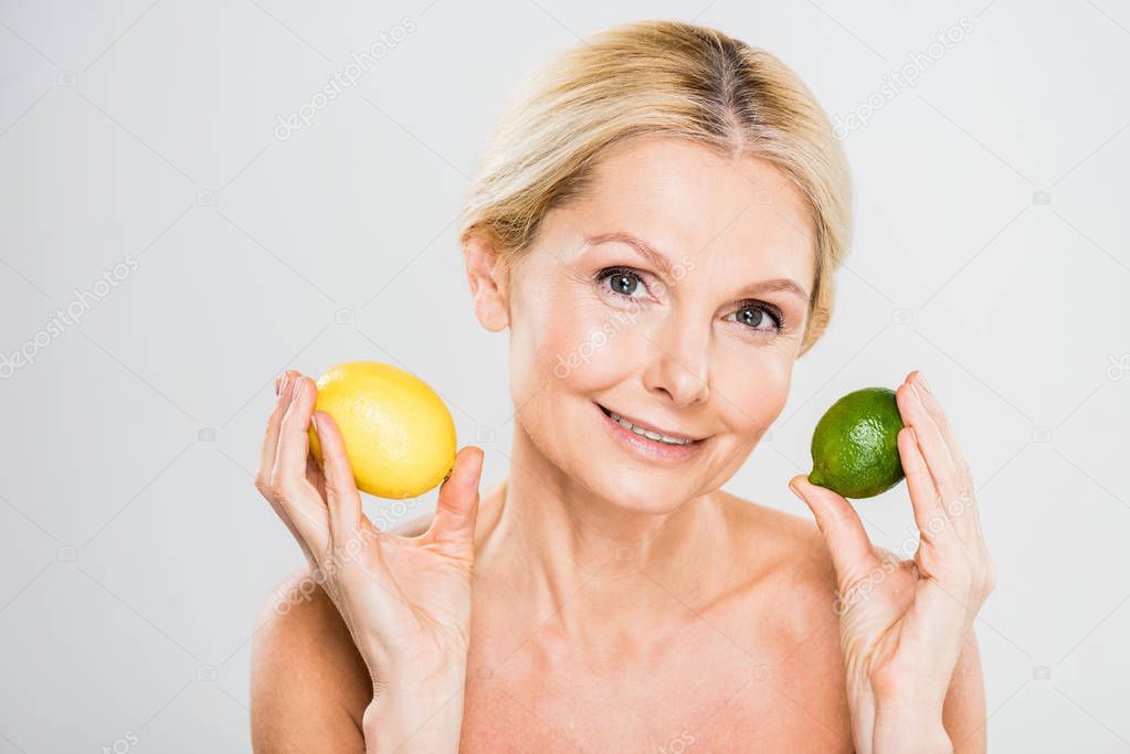 beautiful and smiling mature woman holding ripe lime and lemon and looking at camera on grey background 