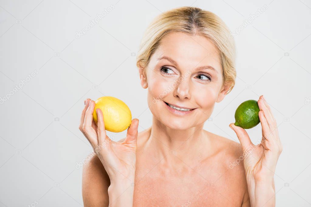 beautiful and smiling mature woman holding ripe lime and lemon and looking away on grey background 