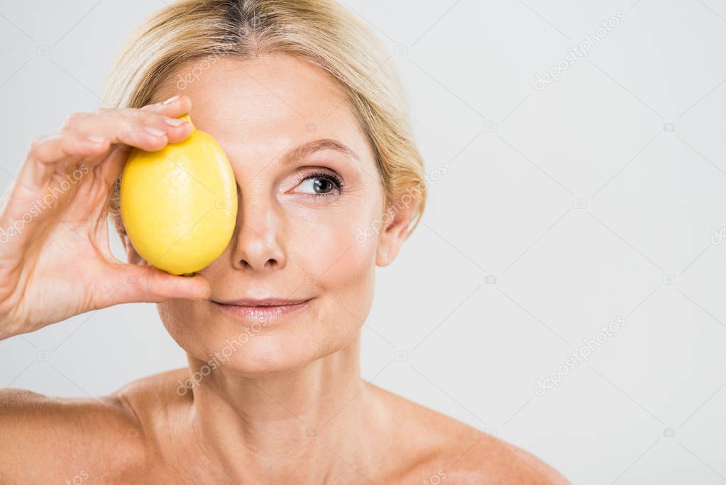 beautiful and mature woman holding ripe lemon and looking away isolated on grey