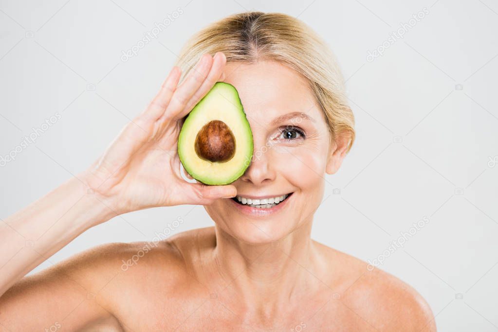 beautiful and mature woman holding avocado and looking at camera isolated on grey