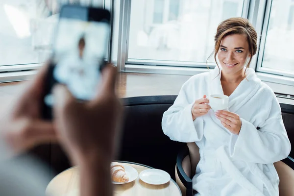 ホテルのバスローブに座りながらカップを保持している魅力的なガール フレンドの写真を撮る男のトリミング ビュー — ストック写真