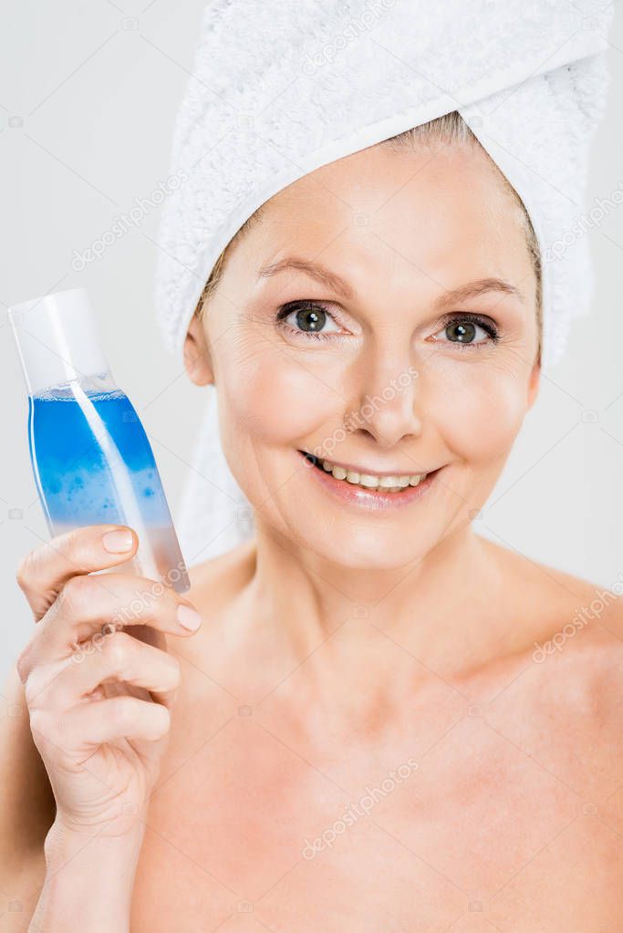 attractive and smiling mature woman in towel holding bottle with micellar water isolated on grey