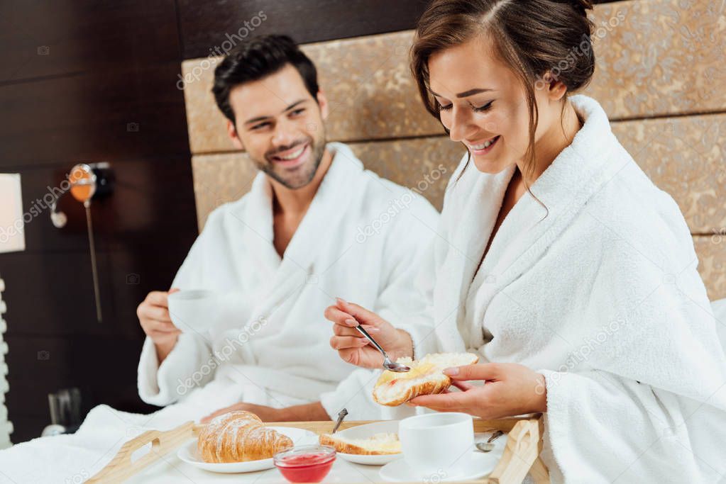cheerful man looking at beautiful woman holding croissant and spoon in bed
