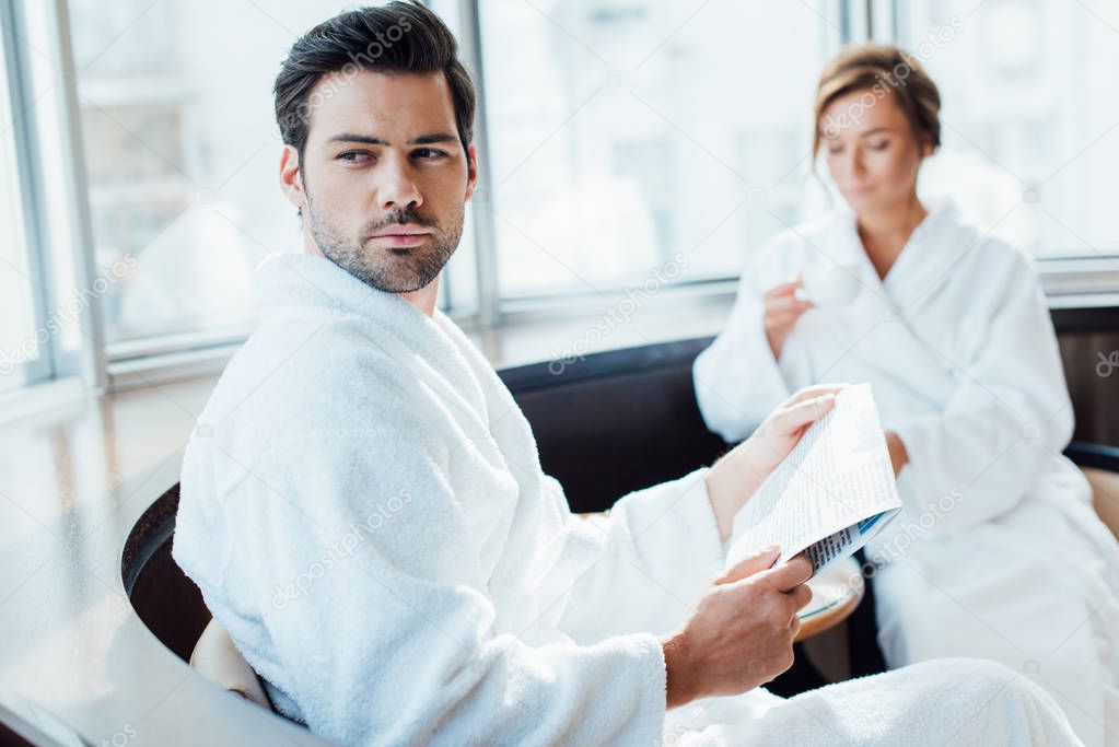selective focus of handsome man holding newspaper in bathrobe near girlfriend with cup