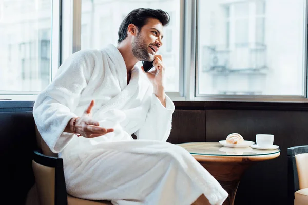 Boldog Szakállas Ember Beszél Smartphone Közelében Reggeli Dohányzóasztal Fehér Fürdőköpeny — Stock Fotó