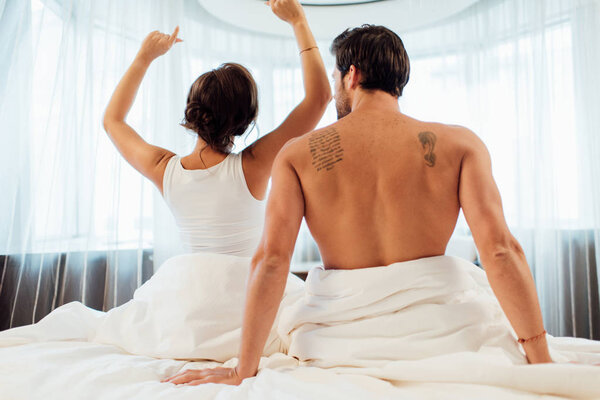 back view of woman stretching in bed near tattooed boyfriend 