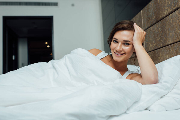 cheerful brunette woman smiling while lying on pillows under blanket in bed 