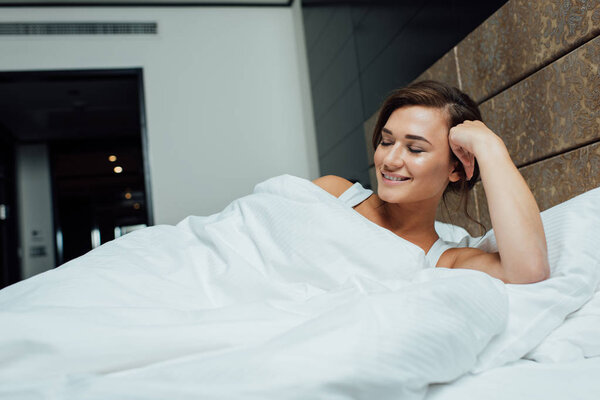 cheerful brunette woman smiling while lying on pillows with closed eyes