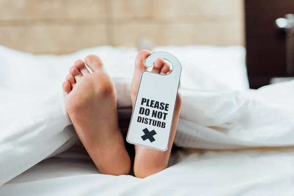 cropped view of woman feet with please do not disturb sign lying in bed 