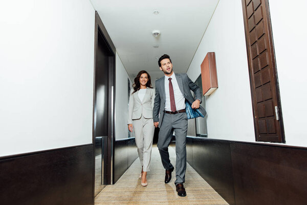 confident businessman and happy businesswoman in formal wear holding hands and walking in hotel corridor 