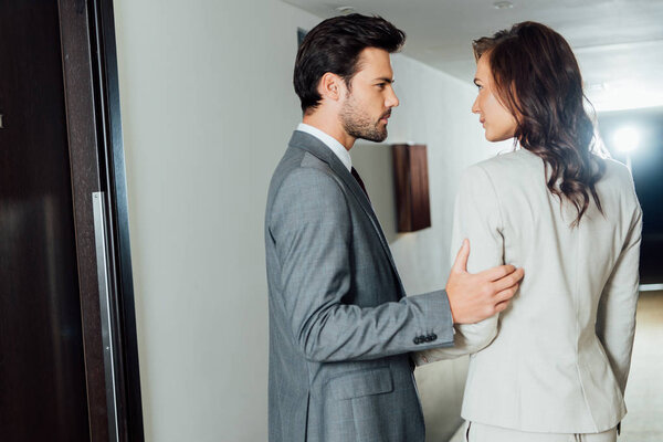 confident businessman touching hand of attractive businesswoman in formal wear while standing in hotel corridor 